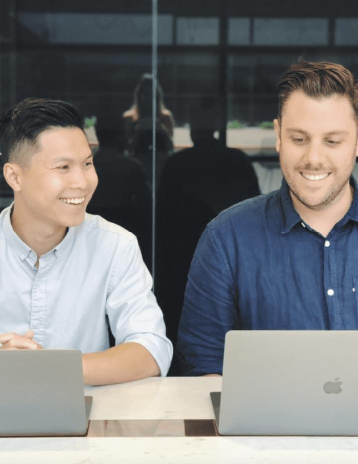 Two men, one asian and one caucasian, smiling and sitting at a table with laptops in a modern office environment. | Future Advisory